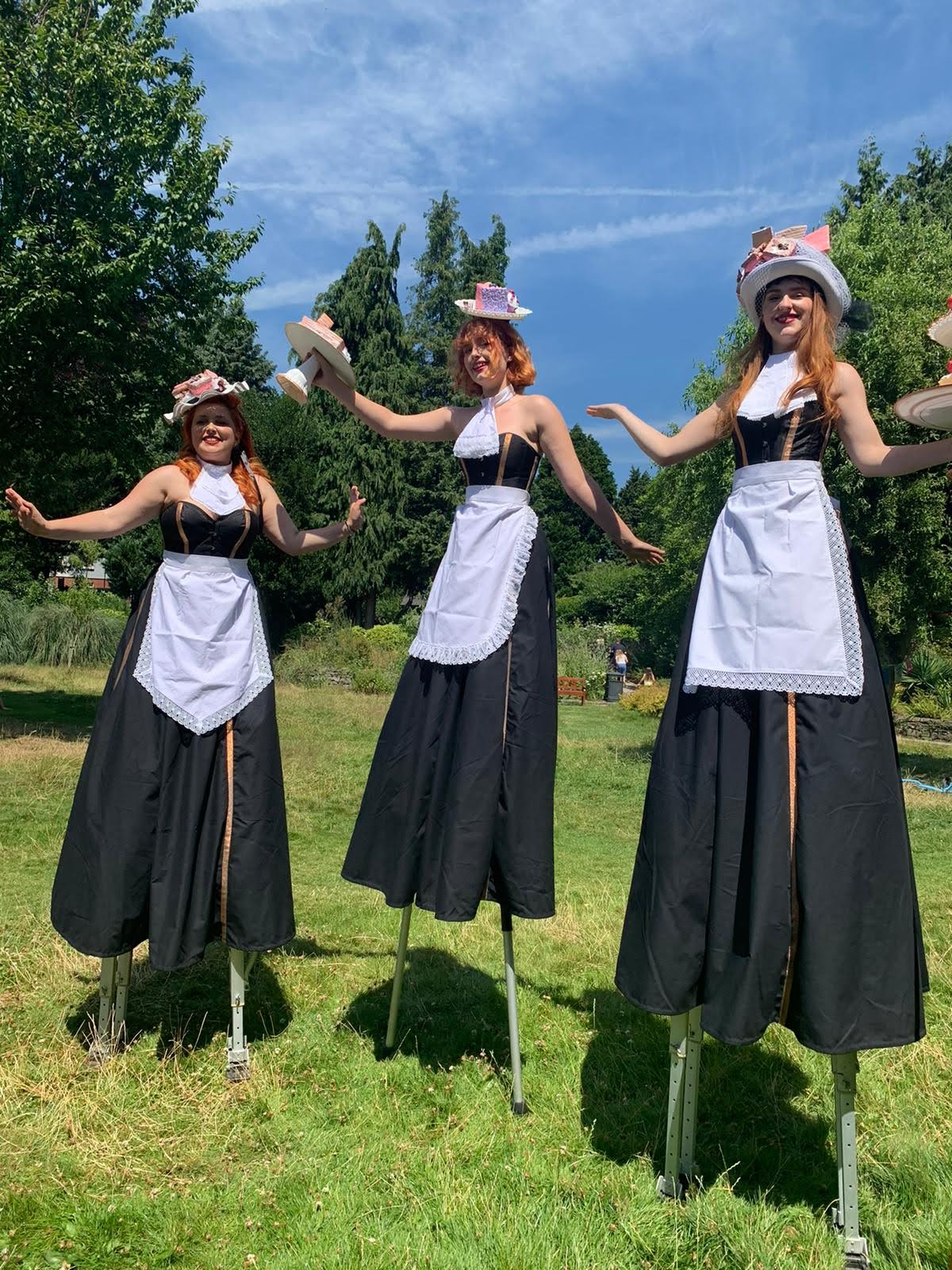 3 women pose on stilts, wearing long black skirts, black corsets, white aprons and hats with cakes on