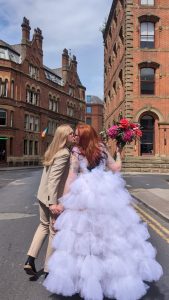 A couple kiss facing away from the camera. One is wearing a multi-tiered white dress made of tulle.